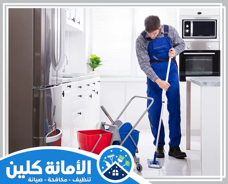 شركة تنظيف فلل بالخبر 0535521559 الامانة كلين عامل نظافة من شركتنا يرتدي زيًا أزرق ويقوم بمسح أرضية مطبخ حديث باستخدام ممسحة وعربة أدوات نظافة احترافية.